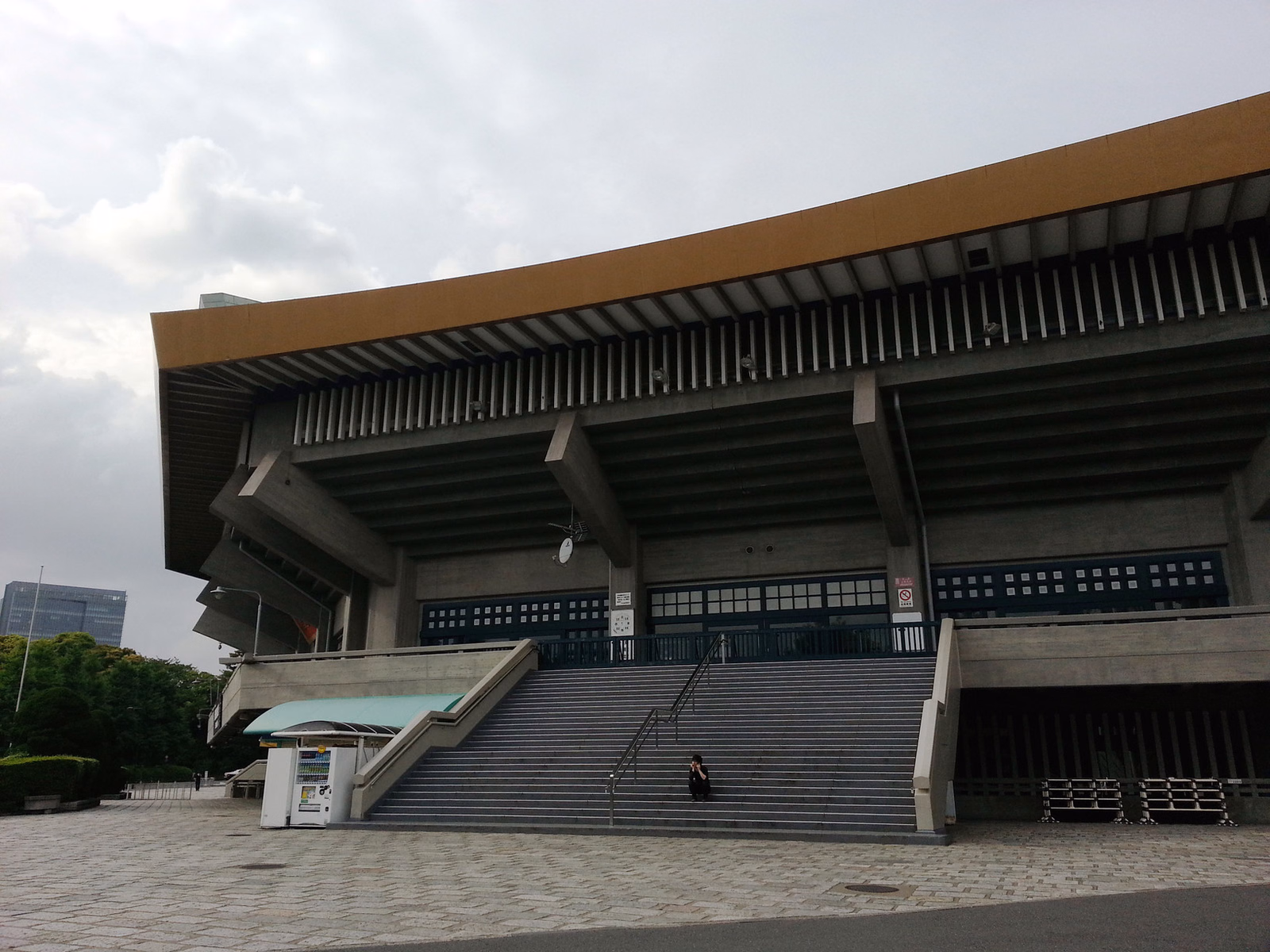 A large modern concert hall with a prominent orange roof and wide exterior staircase is shown under an overcast sky.