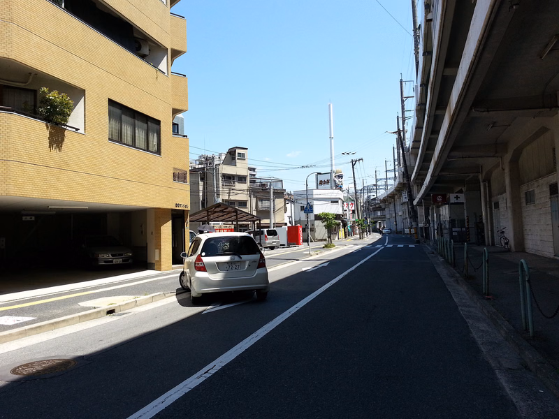 A street scene featuring a modern apartment building on the left and a large concrete overpass on the right, with a white car driving down the road.