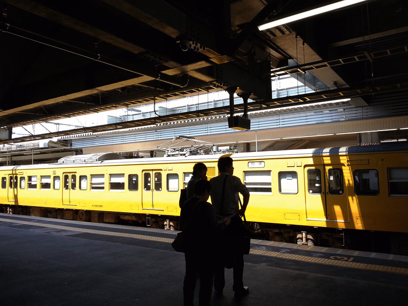 A bright yellow train is stopped at a covered station platform, with several people standing in the foreground.