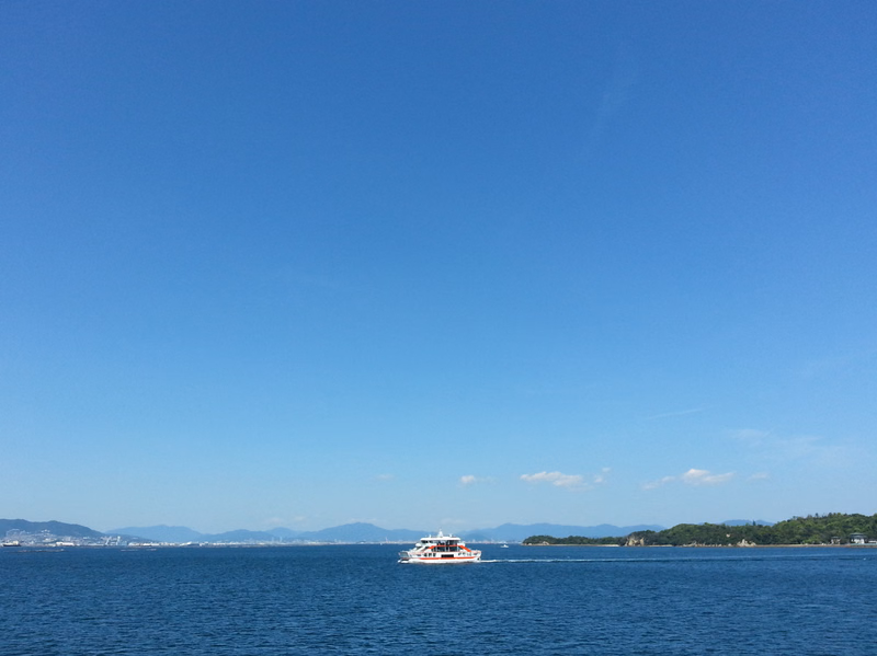 A small passenger boat travels across a vast expanse of blue ocean under a clear, bright blue sky.