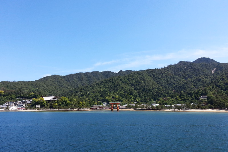A small temple structure stands in the water near a lush, green, forested coastline under a clear blue sky.
