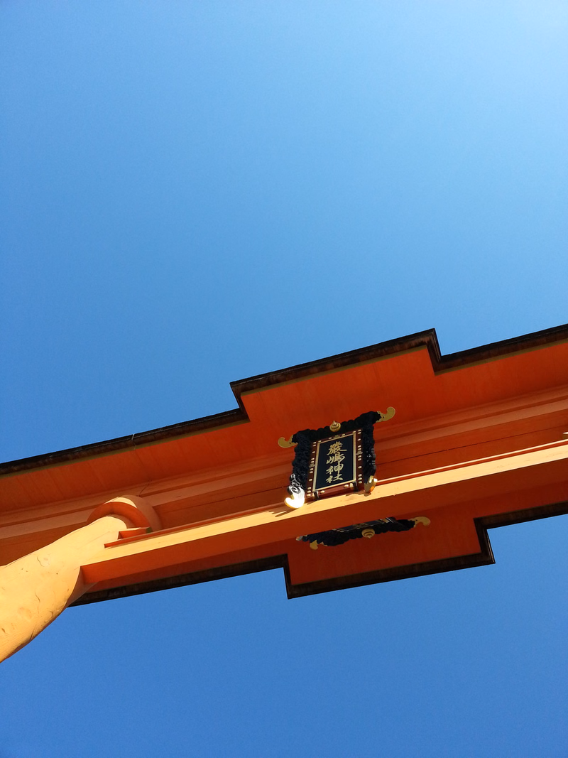 A close-up view of a bright orange torii gate against a clear blue sky, featuring a plaque with Japanese characters.
