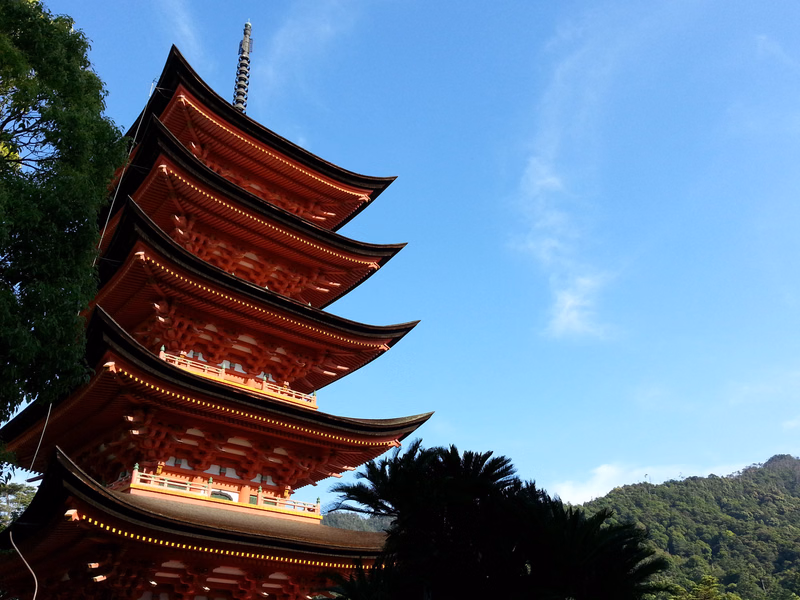 A large, red, multi-tiered pagoda stands against a bright blue sky, with green trees visible in the foreground and background.