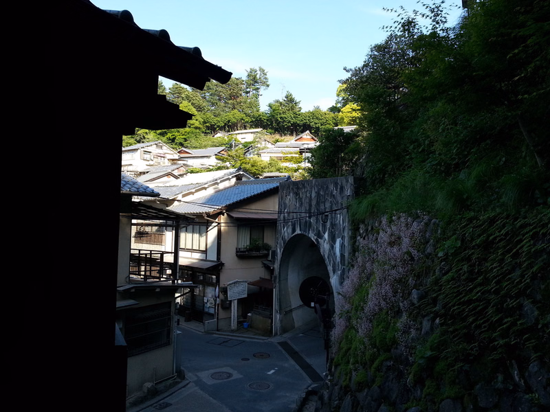A stone archway leads into a traditional town nestled among green foliage on a hillside.