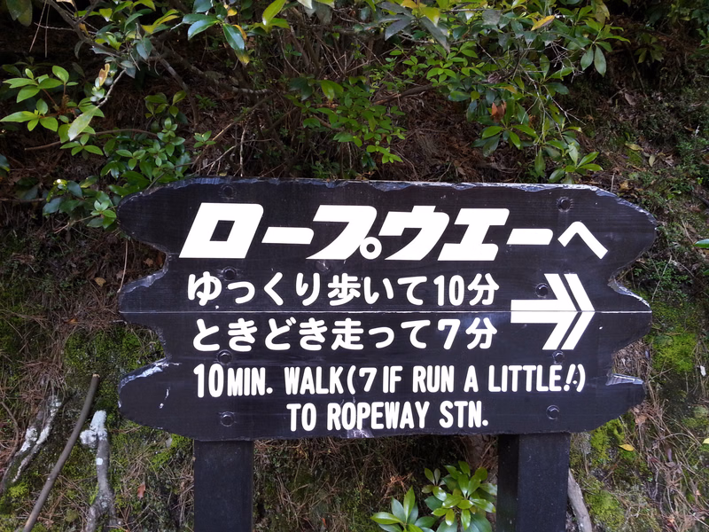 A wooden sign indicates directions to the ropeway with text reading "ロープウェイへ ゆっくり歩いて10分 ときどき走って7分 10MIN. WALK (7 IF RUN A LITTLE!) TO ROPEWAY STN."