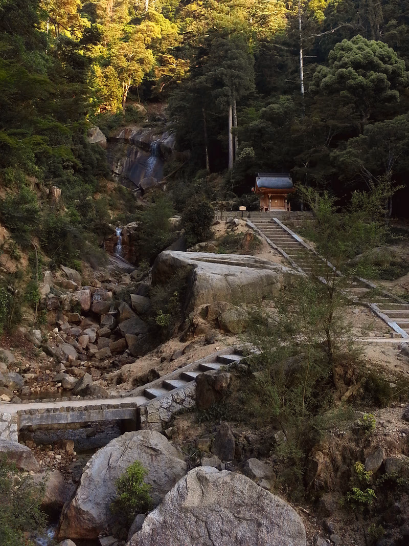 A serene mountain landscape with a waterfall cascading down rocks and a stone path leading to a small wooden building.