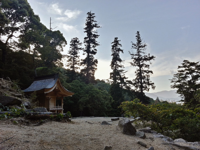 A traditional wooden temple building is nestled among tall trees on a hillside under a cloudy sky.