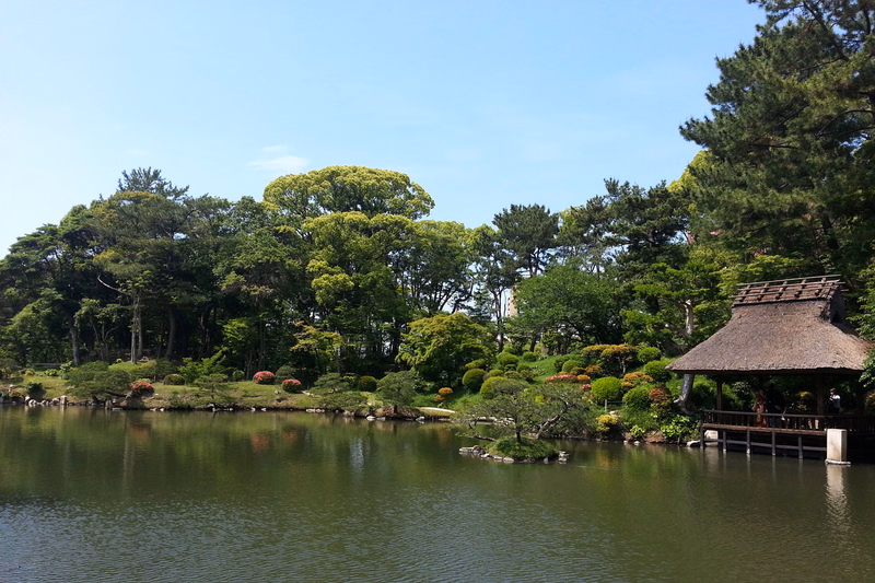 A small thatched gazebo sits beside a calm pond surrounded by lush green trees and manicured garden landscaping under a bright blue sky.