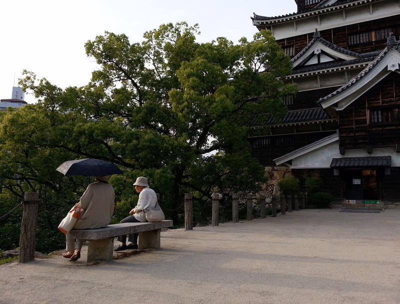 Two people sit under an umbrella on a stone bench near a traditional Japanese temple building surrounded by trees.