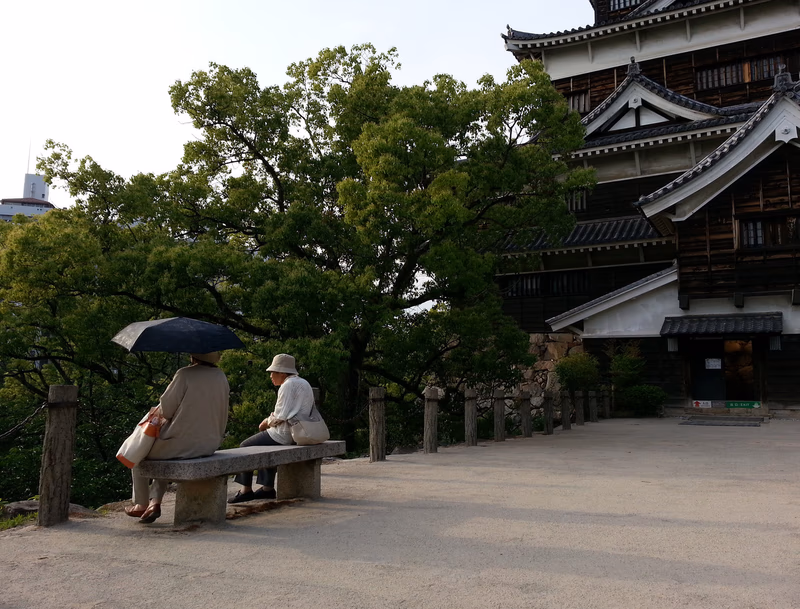 A serene scene of two people sitting under an umbrella near a traditional Japanese building, with a large tree providing shade.