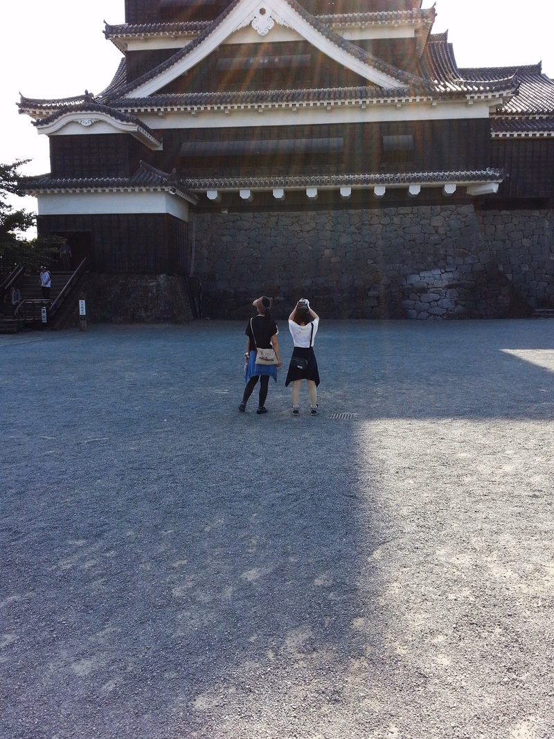 Two women stand in a large gravel courtyard in front of a traditional Japanese castle building.