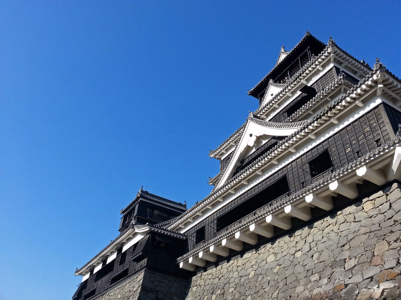 A traditional Japanese castle tower with white plaster walls sits atop a large stone wall against a clear blue sky.