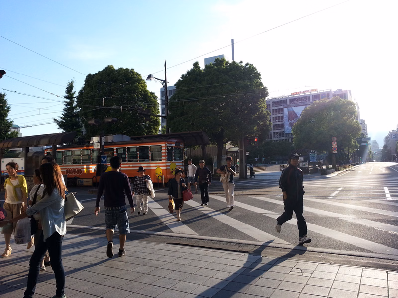 Several people cross a street with a tram visible in the background on a sunny day.