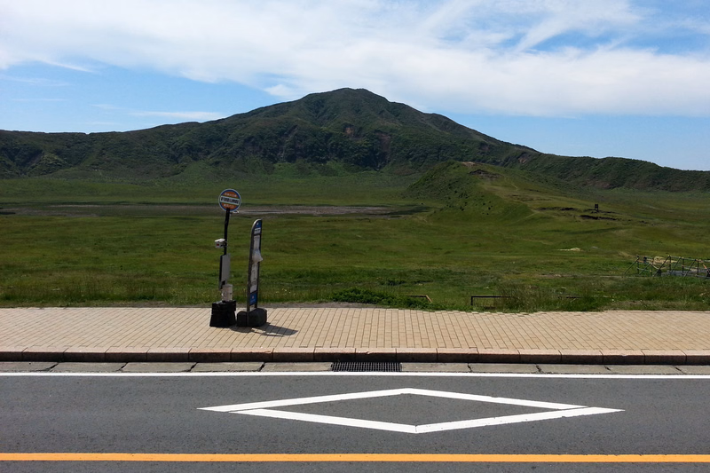 A mountain rises in the distance behind a grassy field, with a road and a circular traffic sign visible in the foreground.