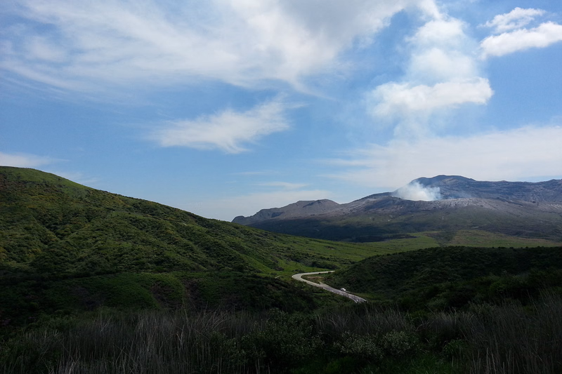A winding road cuts through rolling green hills under a bright blue sky dotted with white clouds, with mountains in the distance.
