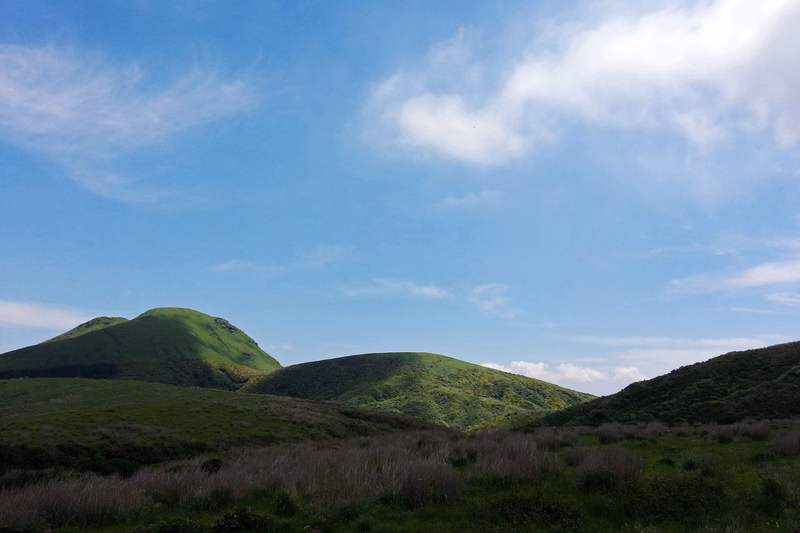 Rolling green hills covered in grass stretch across the landscape under a bright blue sky with scattered white clouds.