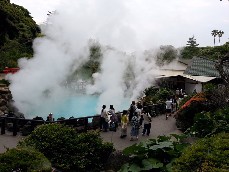A large plume of steam rises from a turquoise hot spring surrounded by lush greenery and buildings, with several people observing.