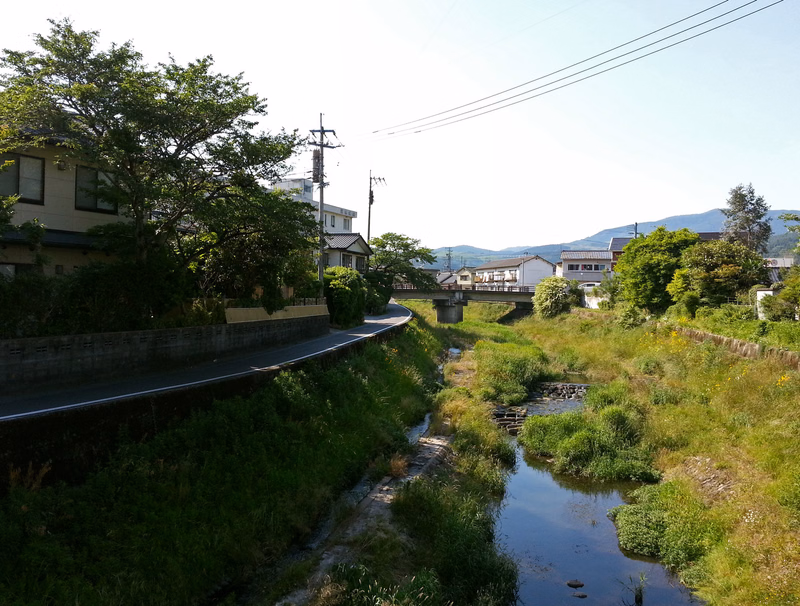 A narrow stream runs alongside a paved road lined with green vegetation and residential houses under a bright sky.