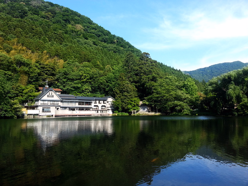 A large white house sits by the edge of a calm lake, backed by a steep, densely forested mountain under a blue sky.