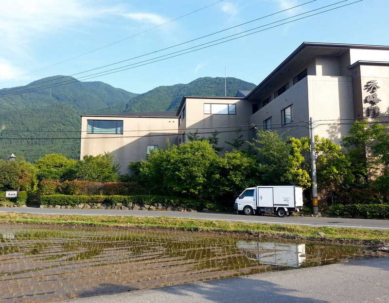 A white truck is parked near a modern building with mountains and rice paddies in the foreground under a blue sky.