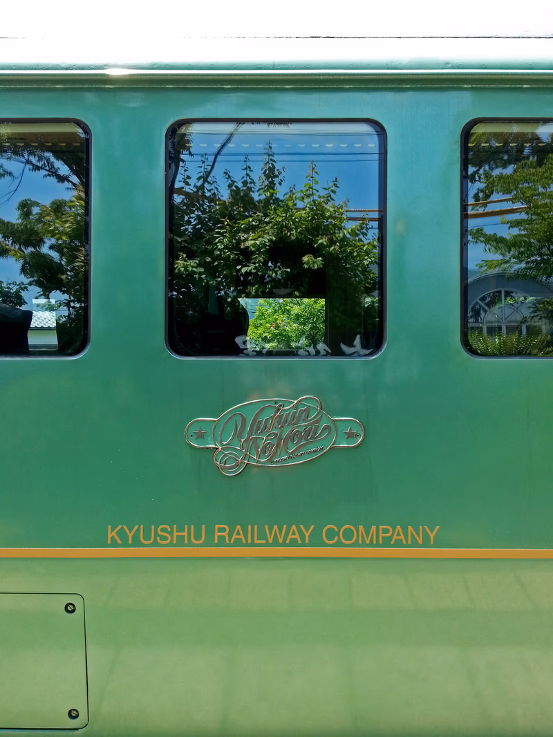 A green train with the name 'Kyushu Railway Company' and the logo 'Yufu' on its side. The train is surrounded by lush greenery and trees.