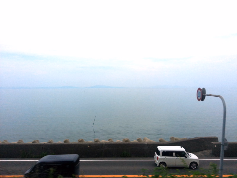 A white car is parked near a road overlooking a vast, calm ocean under a bright, overcast sky.