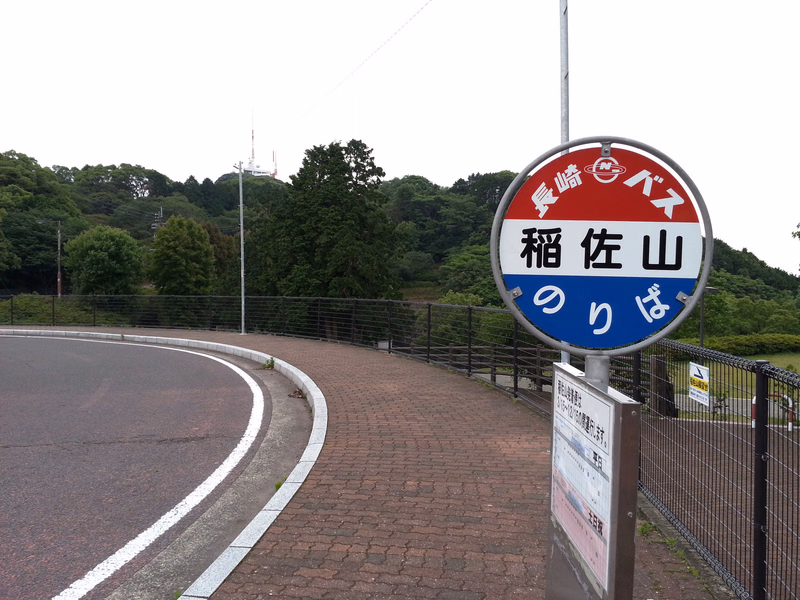 A circular bus stop sign in Japanese reads "長崎バス 稲佐山 のりば" next to a paved road curving past green trees.