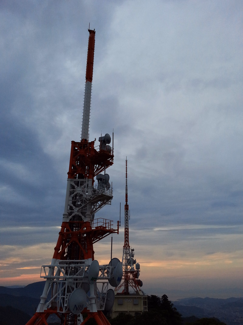 A tall red and white cell tower with multiple antennas stands against a cloudy, twilight sky.