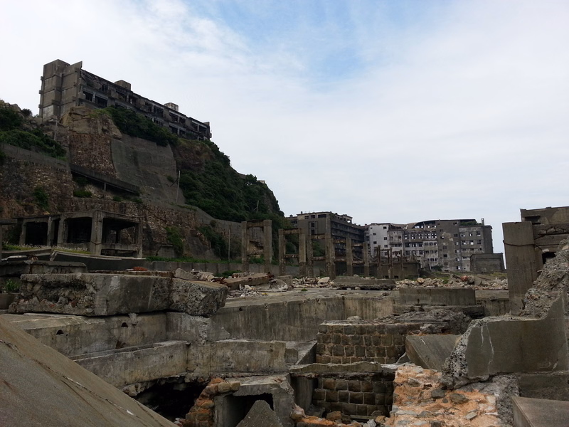Dilapidated concrete ruins and old buildings are situated on a steep hillside under a cloudy sky.