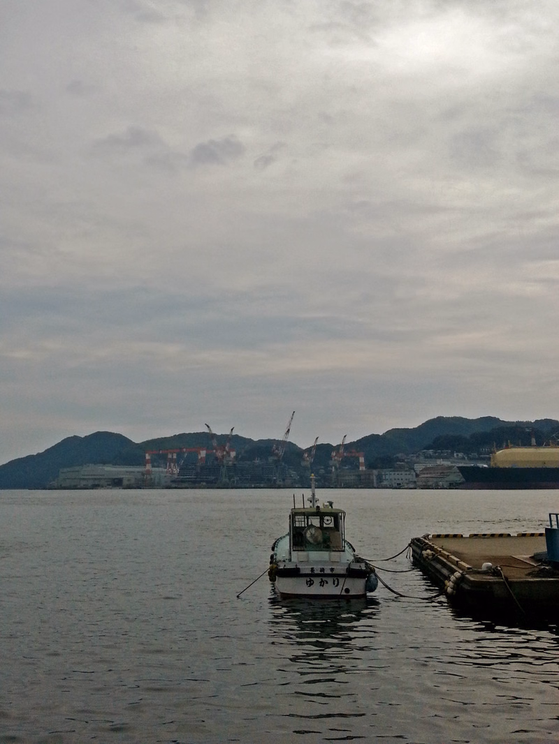 A small fishing boat labeled "ゆかり" floats on calm water near an industrial area under a cloudy sky.