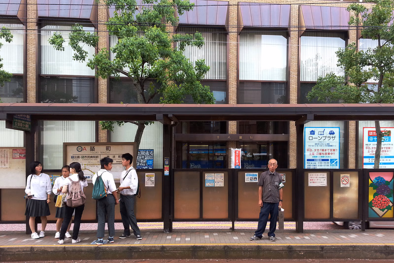 Several people stand in front of a modern commercial building with storefronts, one sign reads "ロンプラザ".