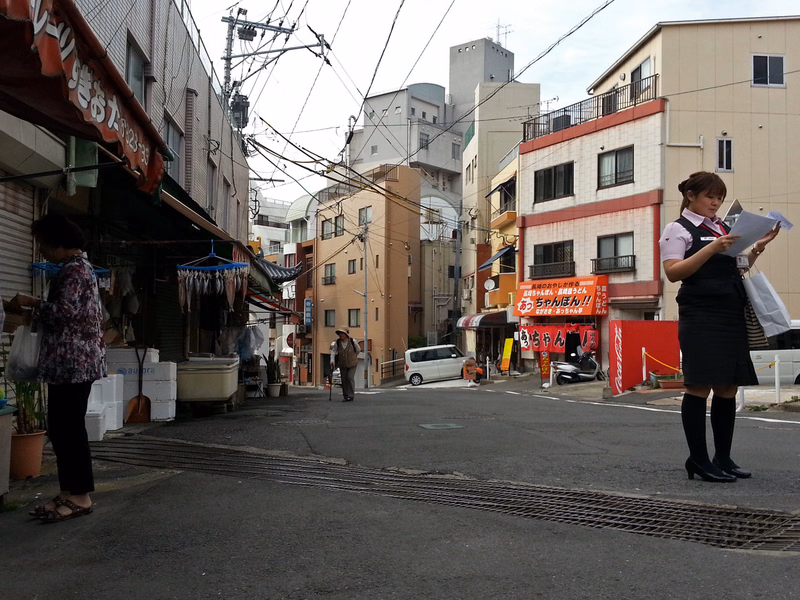 A street scene featuring various buildings, utility poles, and a person holding papers near a shop with a sign that reads "おっちゃん".