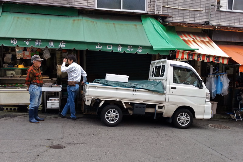 A white pickup truck is parked in front of a vegetable stand with green awnings, and two men are standing near the truck.