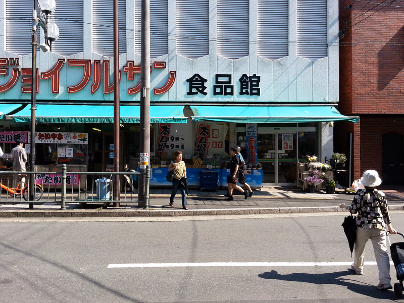 A storefront with a sign reading "ジョイフルサン食品館" is visible on a street with several pedestrians walking by.