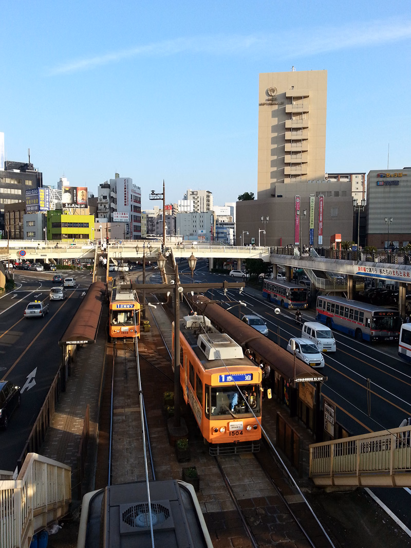 A streetcar is visible on tracks in a busy urban intersection surrounded by various buildings and traffic.
