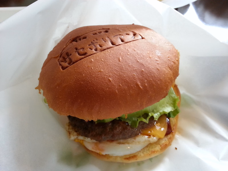 A hamburger with a sesame seed bun, meat patty, fried egg, and cheese is served on white paper.
