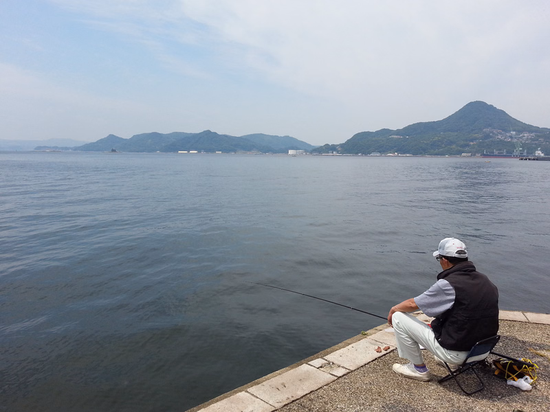 A fisherman on a concrete pier casts a line into the calm ocean water with mountains visible in the background under a bright sky.