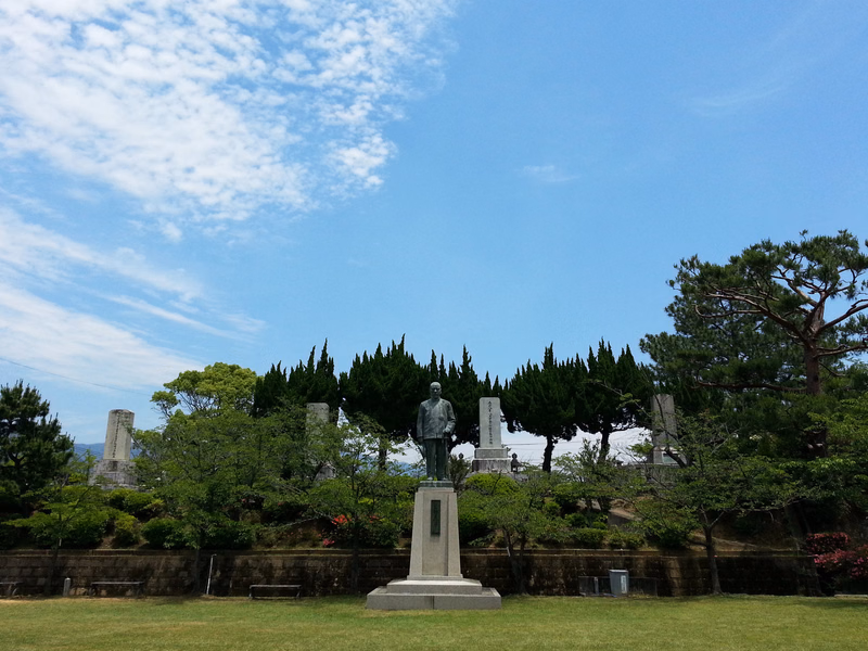 A bronze statue of a man stands on a pedestal in a grassy park area under a bright blue sky with white clouds.