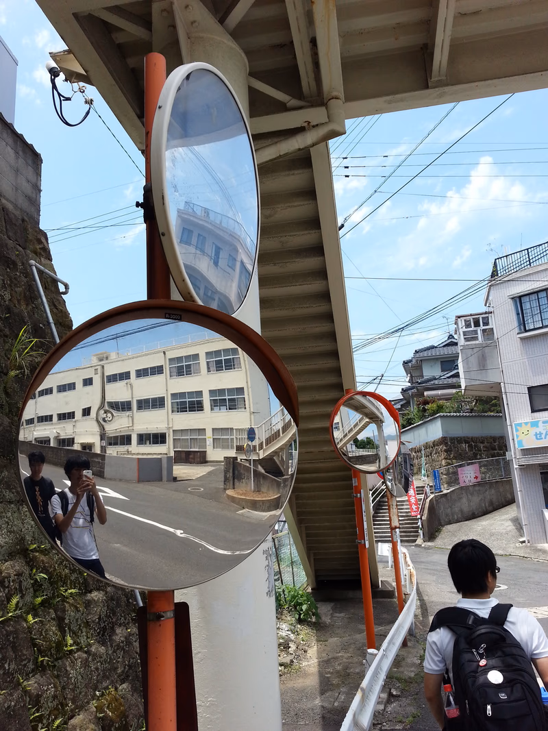 A man is walking down a street under a bridge, and he is looking at a round mirror that reflects a building and a person.