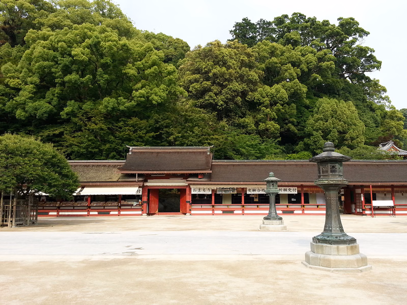 A traditional Japanese temple building with a tiled roof is surrounded by lush green trees, and a stone lantern stands in the foreground.