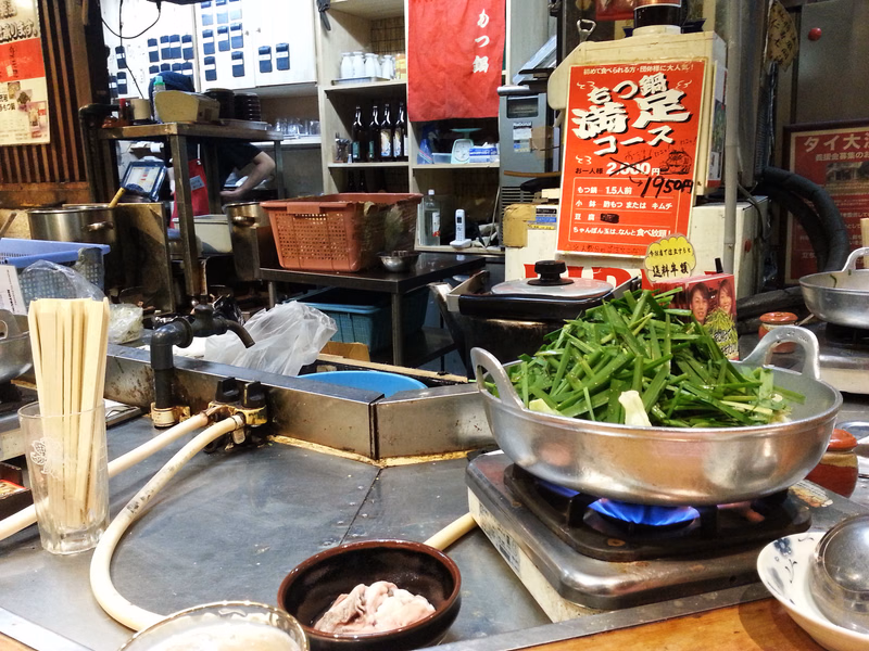 A metal pot filled with green vegetables sits on a small burner in a kitchen setting, next to a sign that reads "もつ鍋 満庭コース 2,950円".