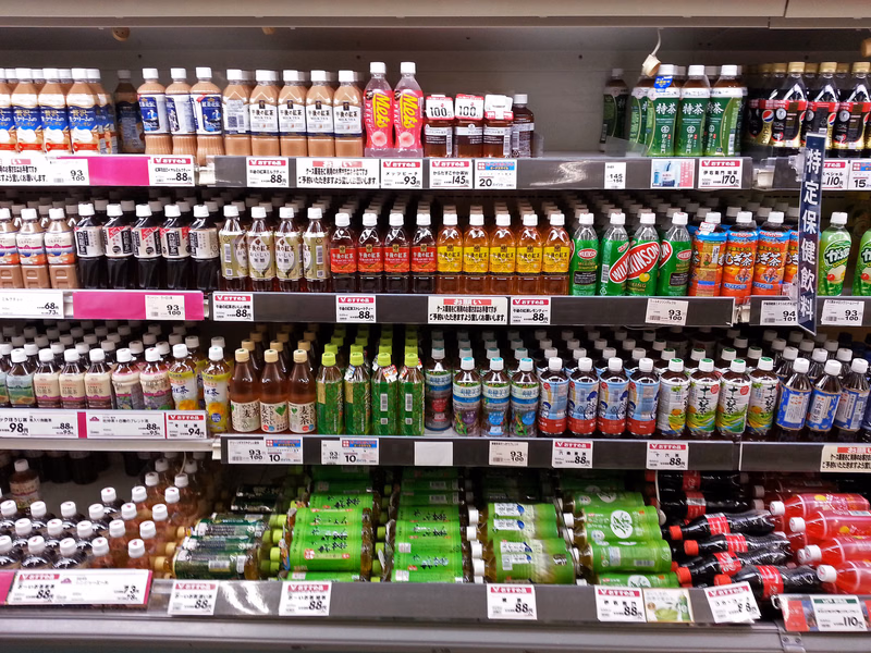 A brightly lit store aisle displays numerous shelves stocked with various brands of soda, juice, and bottled beverages.