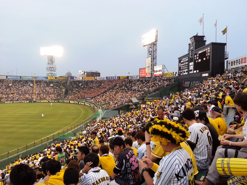 A large crowd fills the stands of a baseball stadium during a game, with players visible on the green field.