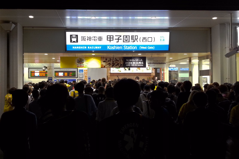 A busy indoor scene at Hanshin Railway Koshien Station (West Gate), showing a large crowd under a station sign.