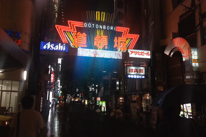 A vibrant night scene in Dotonbori featuring large illuminated signs, including one that reads "DOTONBORI" and another with "Asahi".