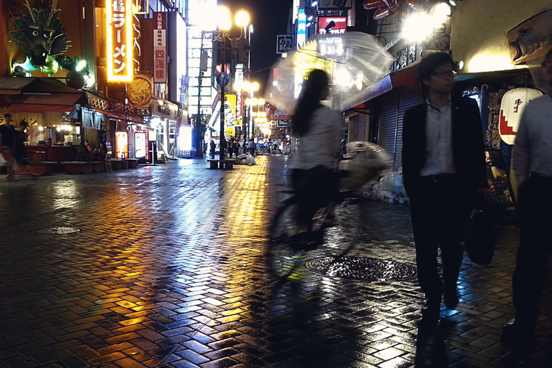 A night in the city, a bustling street, a woman and a man walking, a bicycle, a neon sign, a wet street reflecting lights, and a cityscape at night.