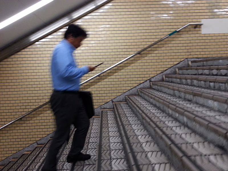A man in a blue shirt is walking down a staircase while using his phone. The scene is set in a subway station with a brick wall and metal handrail.