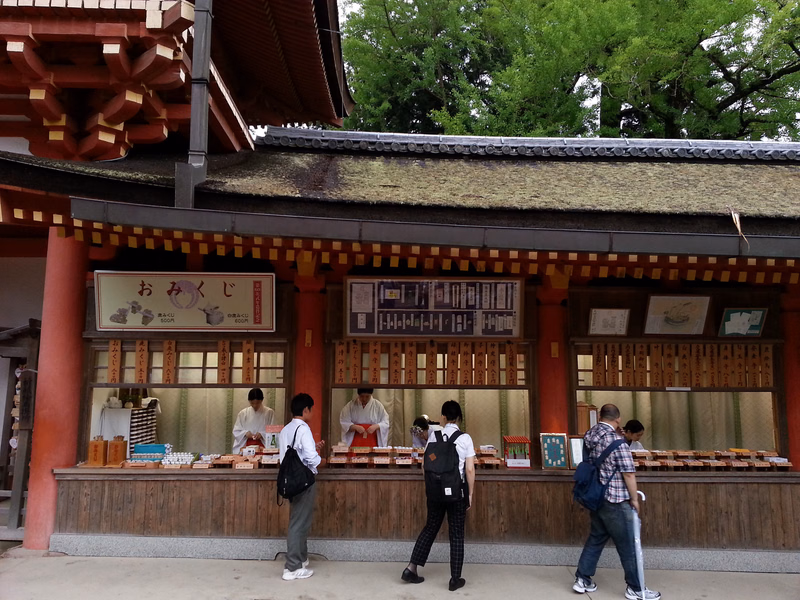 A Japanese shop with a thatched roof displays a sign that reads "おみくじ" and has several people standing outside.