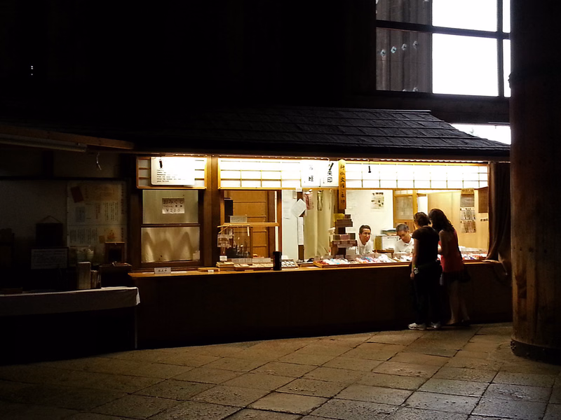 A traditional Japanese tea house with a wooden counter is visible inside a dimly lit building, with bright light coming from large windows.