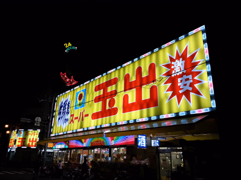 A large, brightly lit yellow and red sign for a supermarket reads "スーパー王出" and features a 24 hour logo.